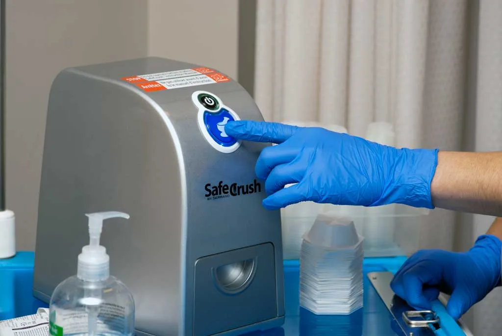A gloved finger pressing the SafeCrush button on a medication cart