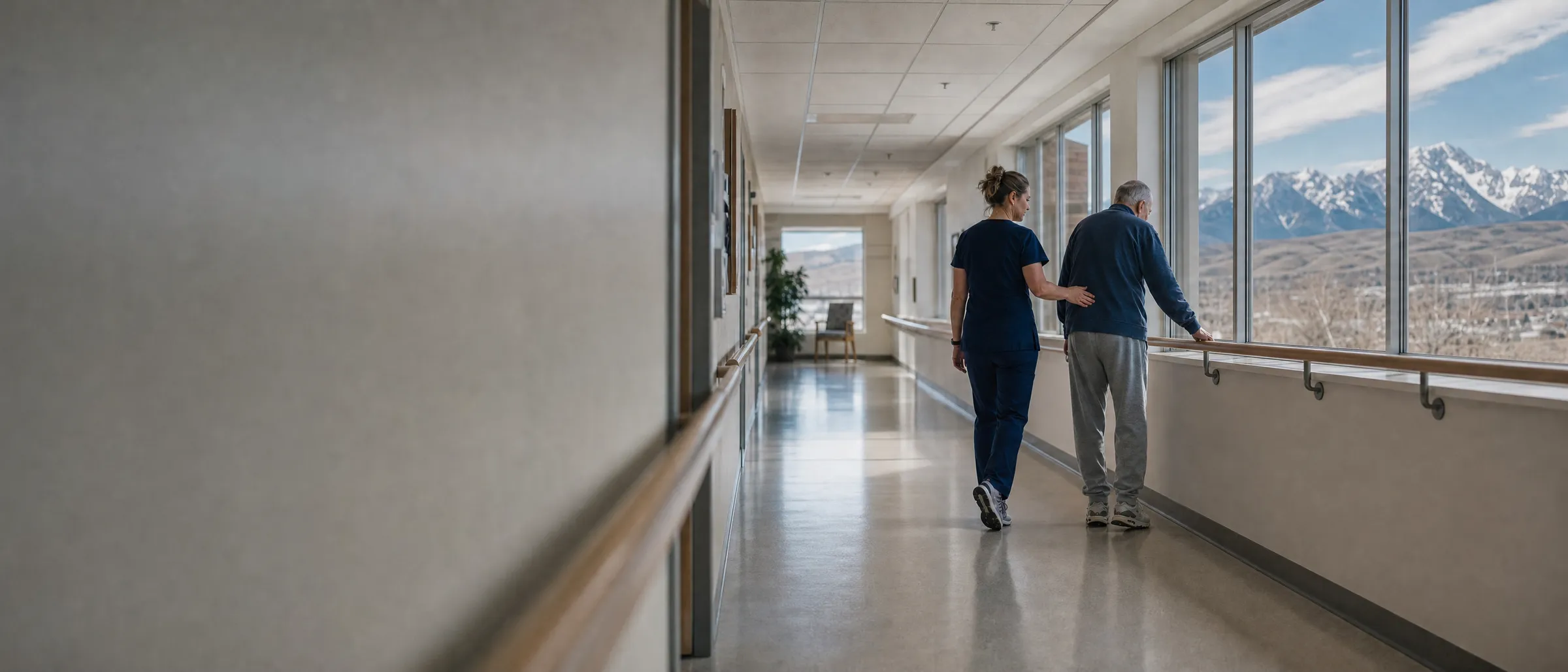 Vue de dos, une physiothérapeute marche à côté d’un homme âgé qui tient la main courante d’un corridor lumineux, avec les Rocheuses visibles par les fenêtres