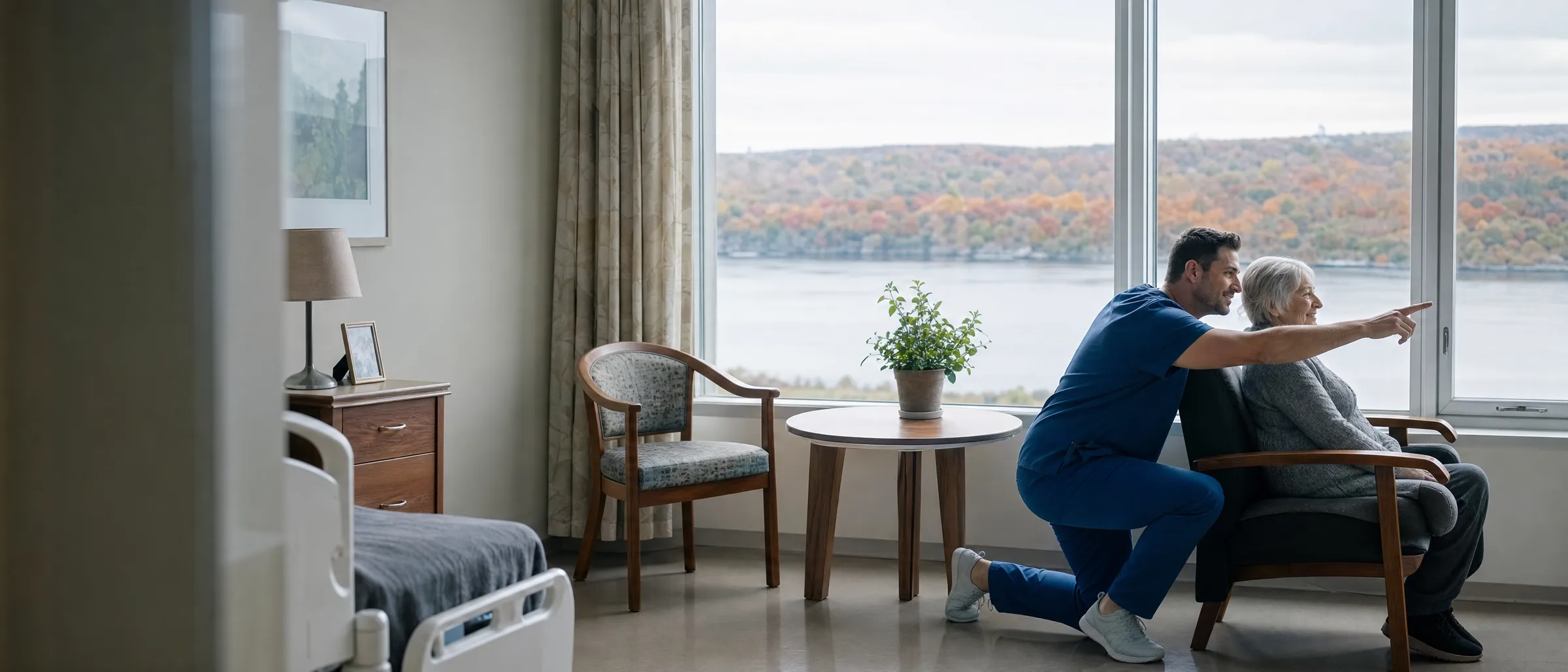 A nurse kneeling beside a resident by a window overlooking a river and autumn hills