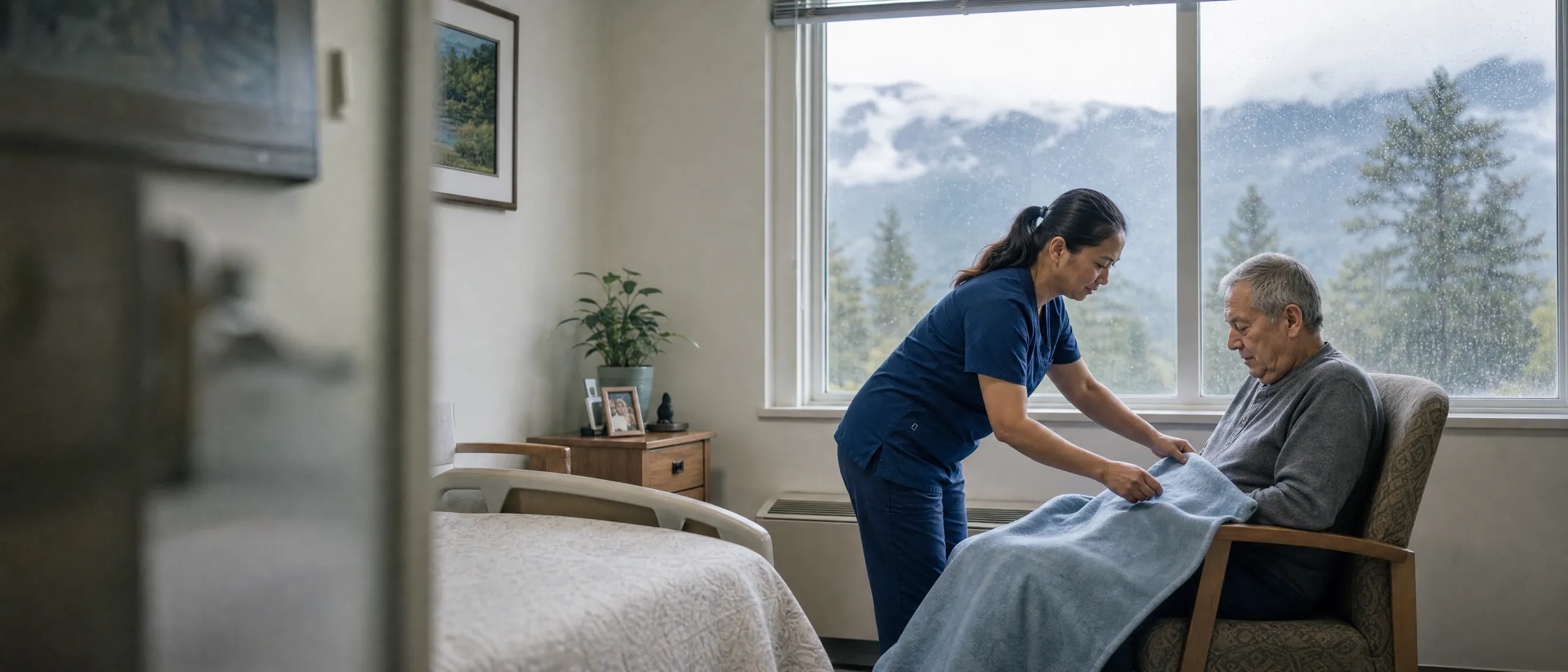 A nurse tucking a blanket over the knees of a resident by a rainy window with evergreens and mountains